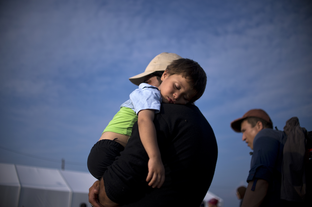 A migrant man holds his sleeping child as migrants and refugees queue to be registered at a camp after crossing the Macedonian-Greek border near Gevgelija on September 23, 2015. European Union leaders hold an emergency migration summit on September 23 amid a growing east-west split after ministers forced through a controversial deal to relocate 120,000 refugees. AFP PHOTO / NIKOLAY DOYCHINOV