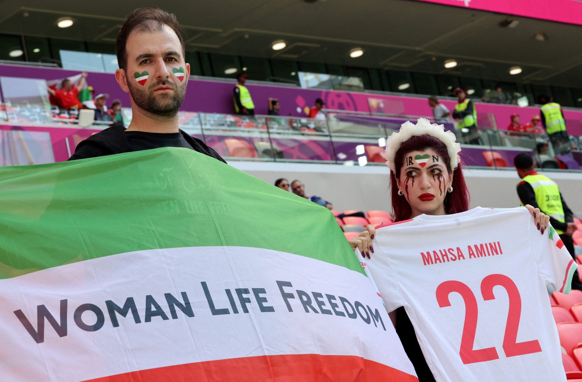 Két rezsimellenes iráni szurkoló az Ahmad Bin Ali Stadionban Irán Wales elleni meccse előtt. Balra a férfi a tüntetések szlogenjével - Nő, Élet, Szabadság - díszített zászlót, jobbra a nő a meggyilkolt Mahsza Amini nevével feliratozott iráni focimezt tart fel.