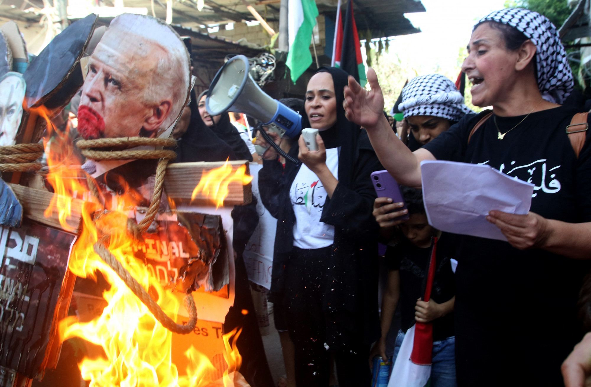 Egy nő beszél megafonba, miközben Joe Biden amerikai elnök képét égetik egy tüntetésen az Ain al-Hilweh palesztin menekülttáborban, a dél-libanoni Szidón kikötőváros közelében 2023. október 18-án, aznap, amikor az amerikai elnök a szomszédos Izraelbe látogatott, és egy nappal a gázai Al-Ahli al-Arabi kórház elleni, több száz halálos áldozatot követelő támadás után.
