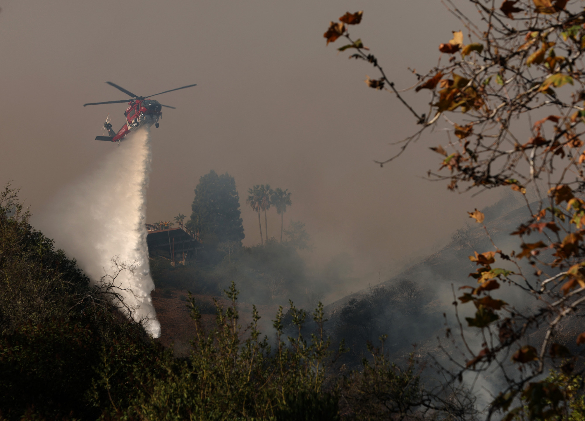 A lángoló vegetációt vízzel bombázó tűzoltó helikopter a kaliforniai Palisades légterében 2025. január 11-én